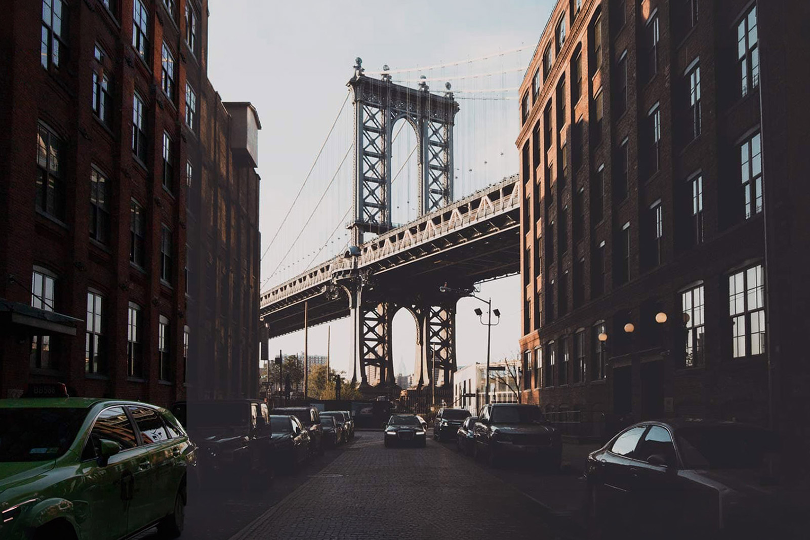 A before-and-after split photo of a cobblestone street in DUMBO, Brooklyn, framed by brick buildings and looking toward the Manhattan Bridge. The edited portion showcases a dark and moody cinematic style with deep shadows, high contrast, and muted tones.