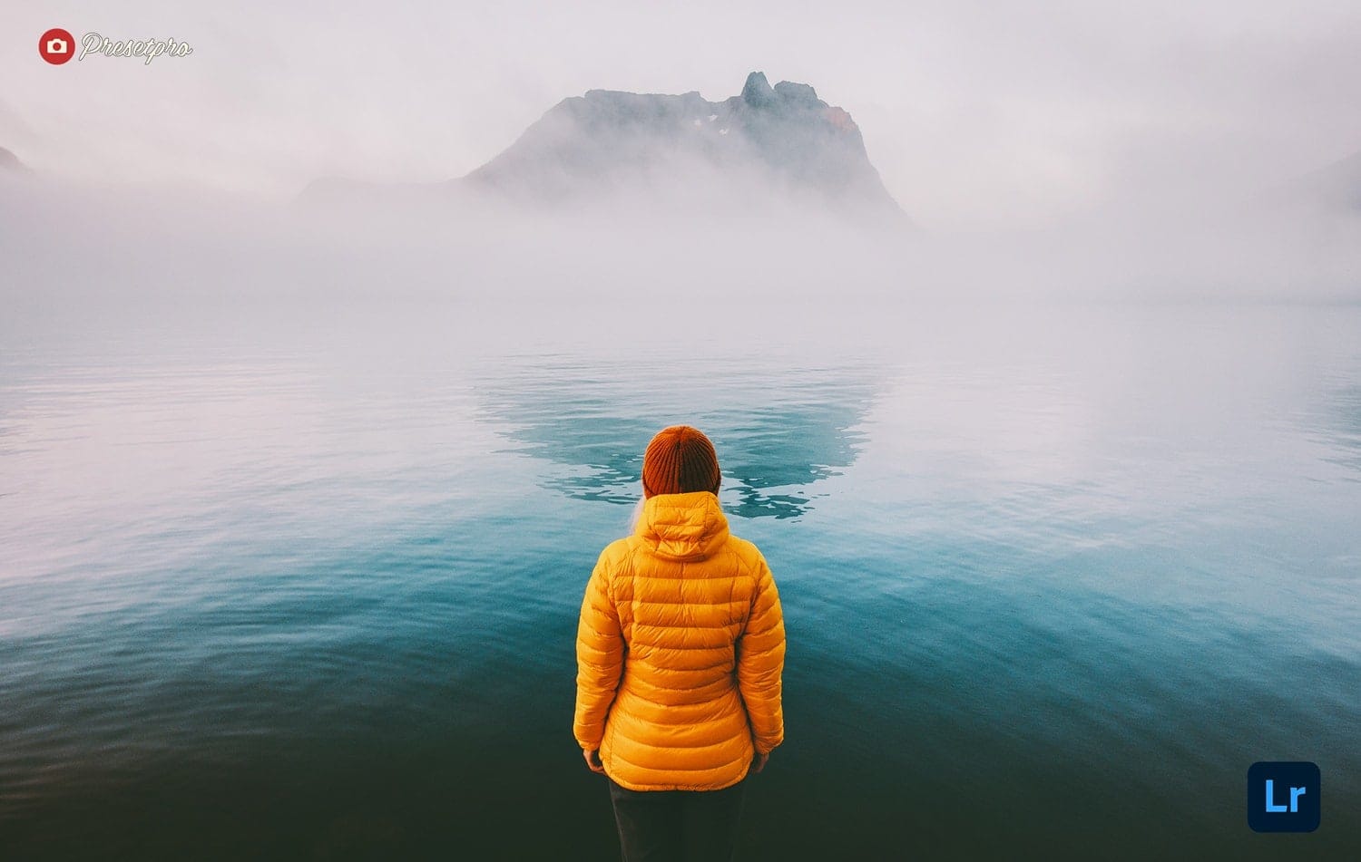 A person in a yellow jacket looking out over a misty lake with mountains fading into fog, edited with the free Moody Lake Lightroom preset and profile by Presetpro.com.